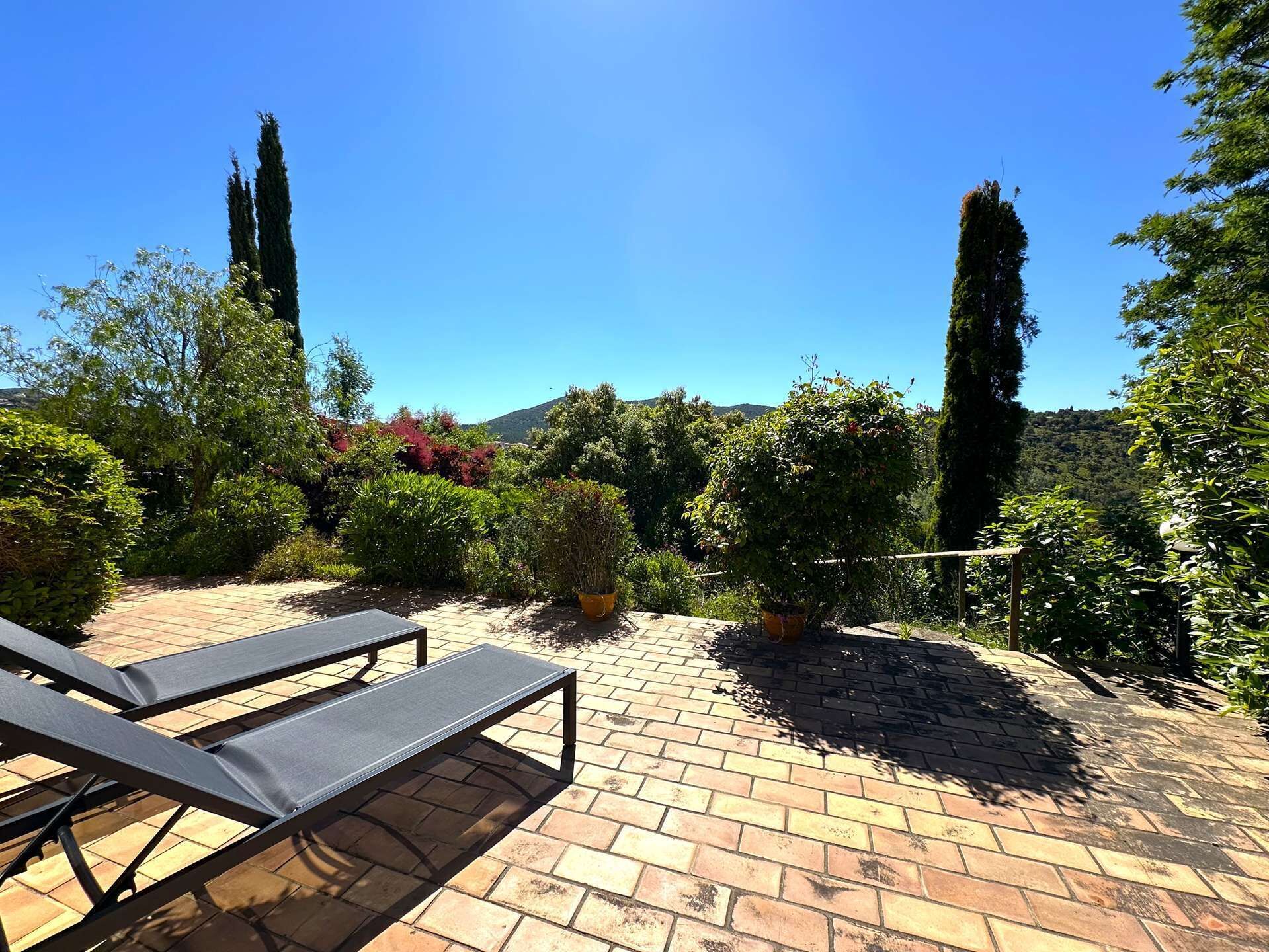 A patio with two lounge chairs, a brick floor, and a variety of potted plants and trees.