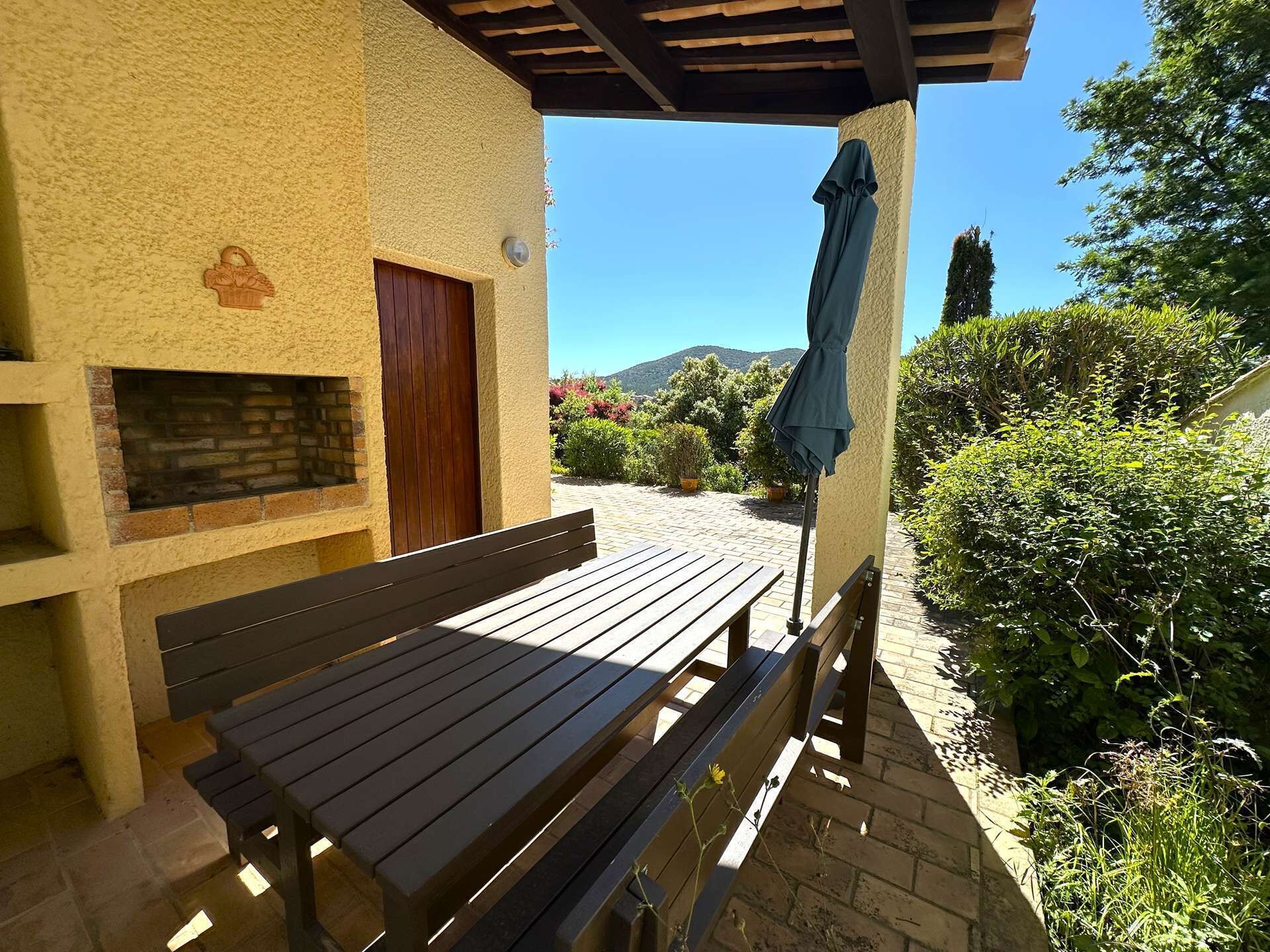 Un banco de madera bajo una pérgola, un muro de ladrillo con una puerta de madera y un cielo azul despejado.