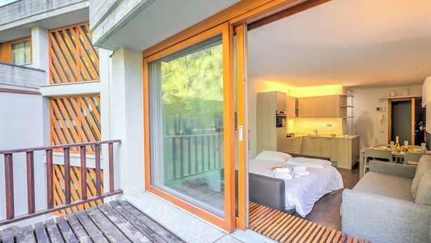 Moderni hotellihuone, jossa on parveke, sänky, sohva, pieni keittotila ja liukuovet, jotka johtavat parvekkeelle.
