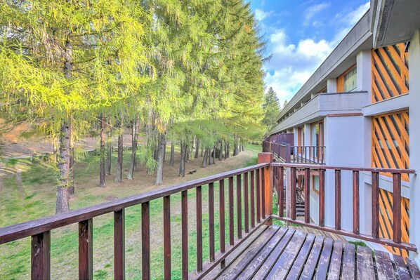 A wooden deck with a railing, a building with a modern design, and a forested area in the background. - Studio La Finestra Sulle Piste With Balcony and Garage (Cesana Torinese)