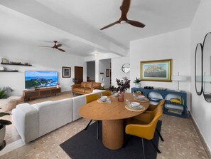 A modern living room with a TV, sofa, dining table, and ceiling fans. - Casa Verde | Historic House For 6 @ San German (San German)