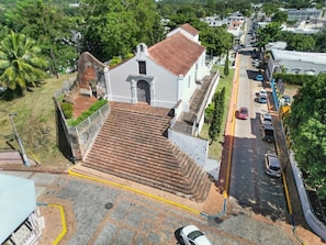 A white building with a tiled roof and a staircase leading to a door. - Casa Verde | Historic House For 6 @ San German (San German)