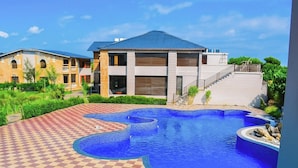 A swimming pool with a checkered pattern around it, a modern building with a blue roof, and a staircase leading to a balcony. - Amaris Greens Resort (Ramnagar)
