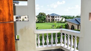 A balcony with a white balustrade, a view of a building with a blue roof, and a green lawn. - Amaris Greens Resort (Ramnagar)