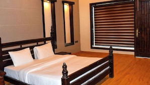 A double bed with a wooden frame and headboard, two bedside lamps, a window with blinds, and a wooden door. - Amaris Greens Resort (Ramnagar)