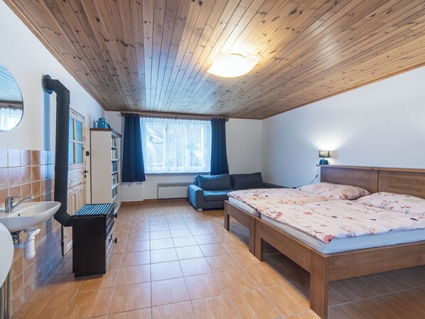 Una camera da letto con soffitto in legno, un letto con biancheria da letto a fiori, un divano, una finestra con tende, un termosifone e un bagno con lavandino e specchio.