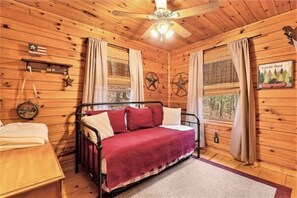 Una habitación tipo cabaña acogedora, con cama, paredes de madera, ventilador de techo y ventana con cortinas.
