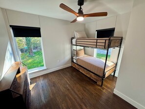 Una habitació amb una llitera, un ventilador de sostre, un escriptori amb calaixos i una finestra amb vista a vegetació.