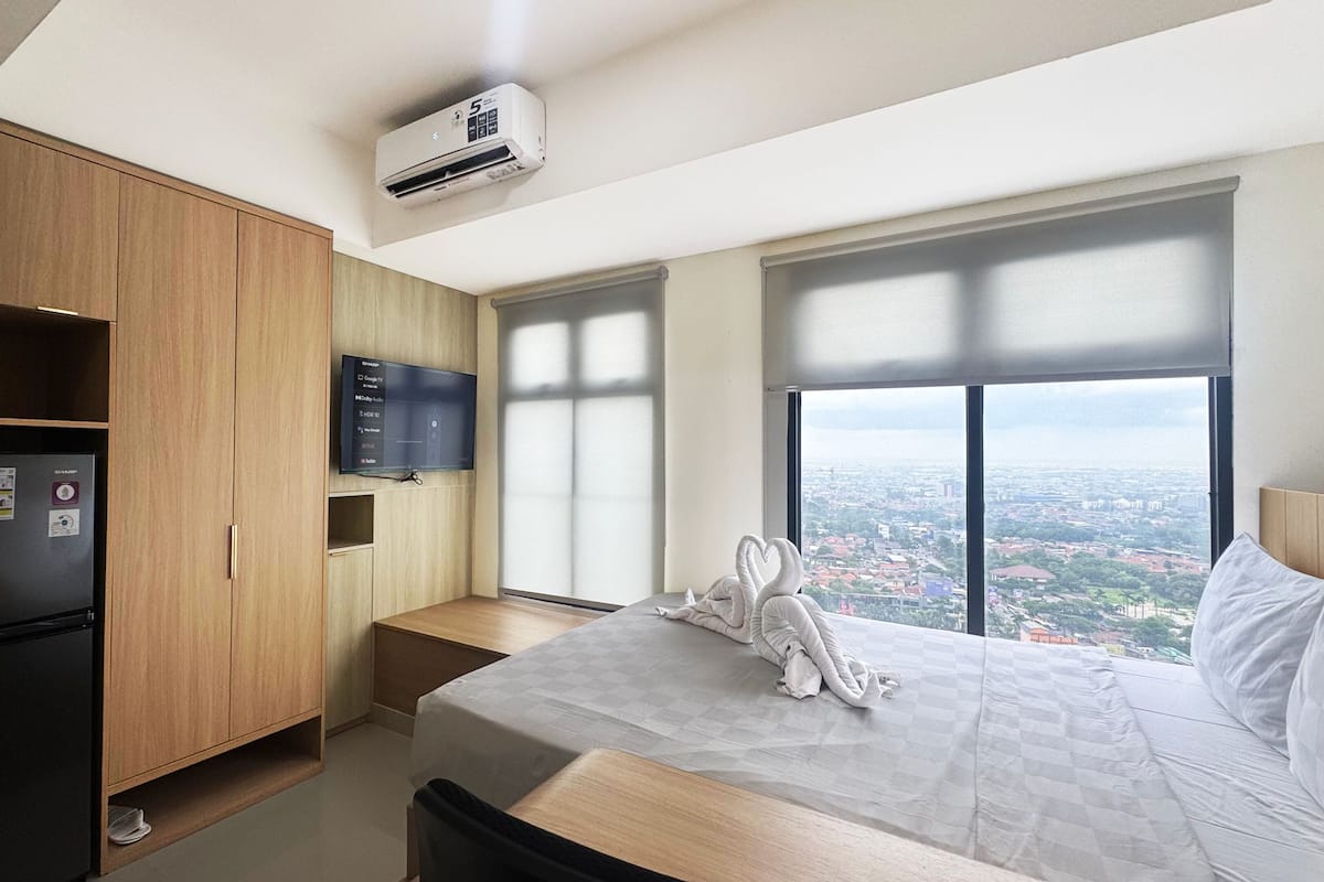 A hotel room with a bed, a minibar, a television, and a large window with a city view.