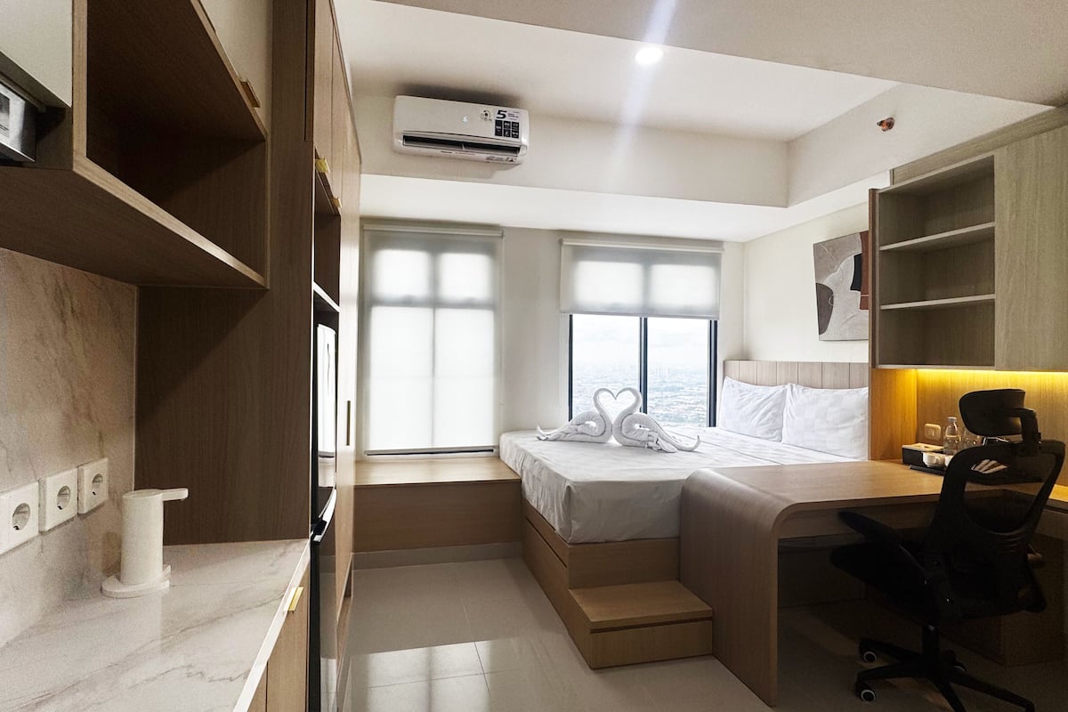 A compact hotel room with a bed, a desk, a chair, a small table, and a window with a city view.