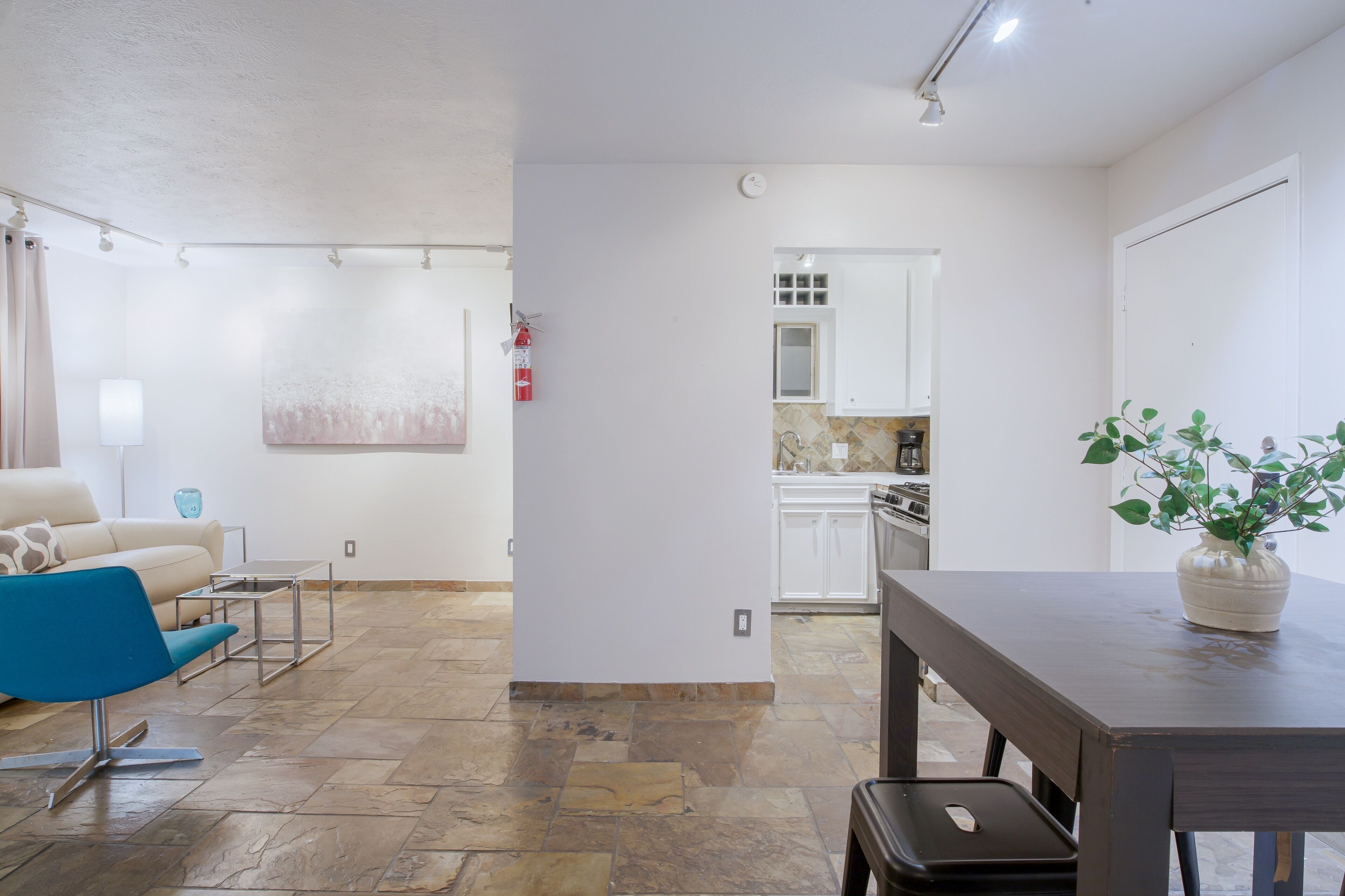 A modern living room with a blue chair, a dining table, and a kitchen area visible through an open door.