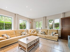 Ein geräumiges Wohnzimmer mit einem beigefarbenen Sofagarnitur, einem Couchtisch und großen Fenstern mit Blick auf Grünflächen.