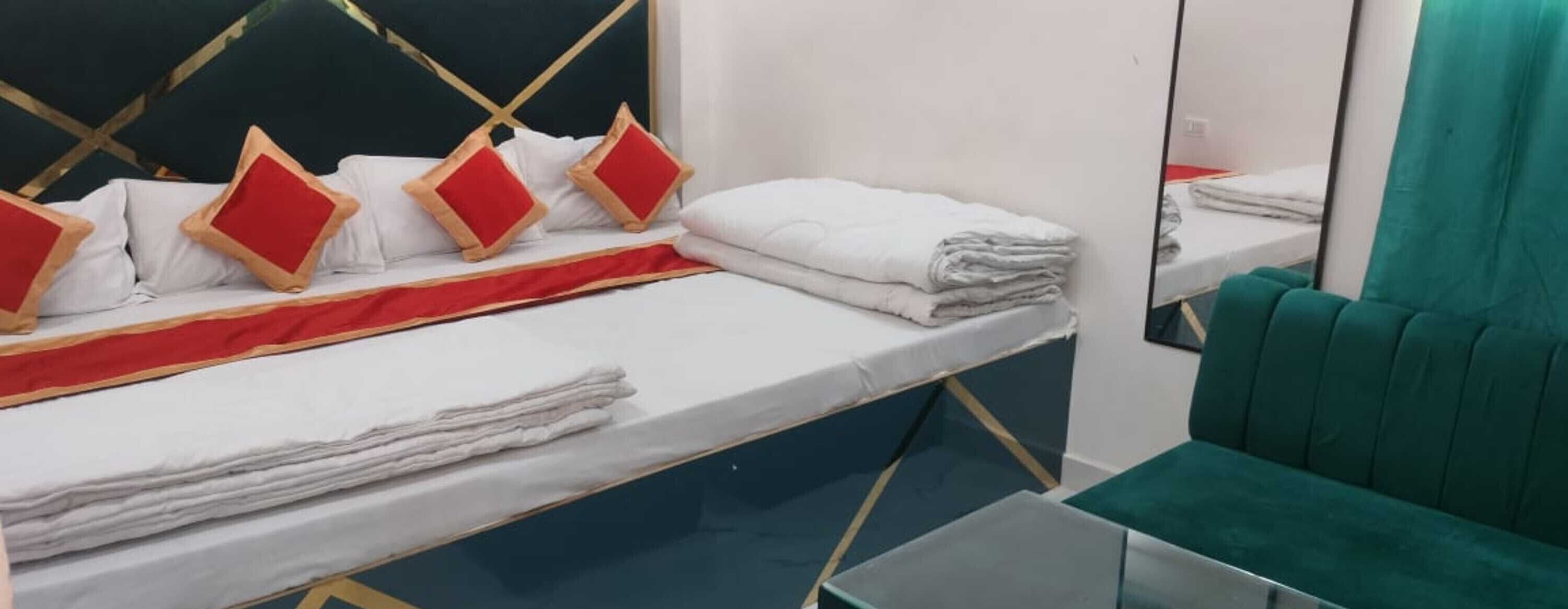 Una cama bien hecha con sábanas blancas y almohadas rojas, una silla color turquesa y un espejo que refleja la habitación.