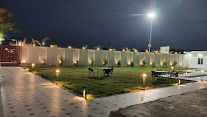 A well-lit outdoor area with a paved walkway, a grassy lawn, and tables and chairs arranged for outdoor dining. - Anandam Hotel (Pindra)