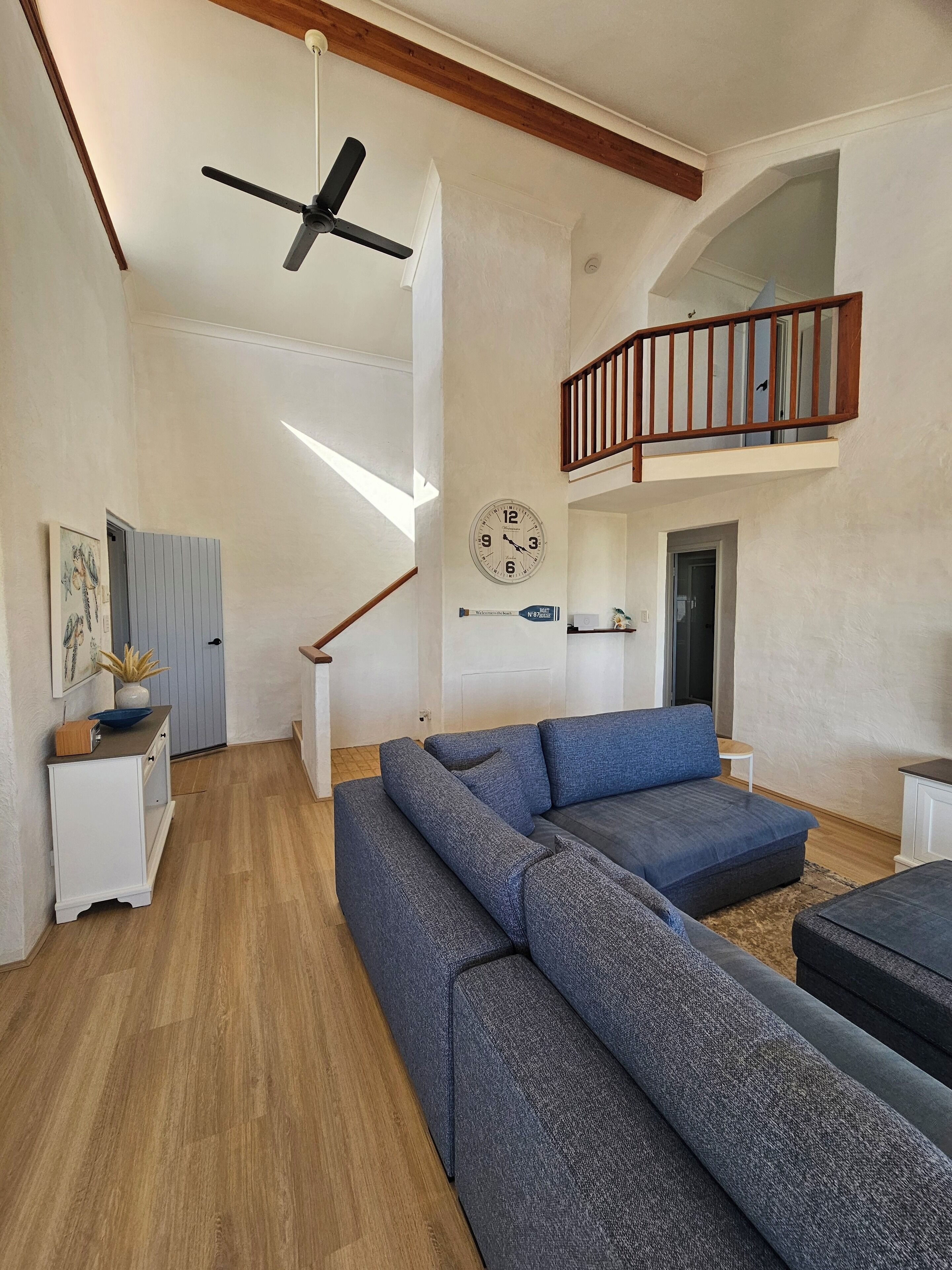 Un salon spacieux avec un grand canapé d’angle bleu, un plancher en bois, un ventilateur de plafond, une horloge et un escalier menant à l’étage supérieur.