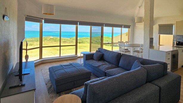 A spacious living area with a large gray sectional sofa, a small round table, and a flat-screen TV on a white stand. A kitchen area is visible in the background with a large window offering a view of the ocean. - Oceanfront retreat with breathtaking views steps from the shore. (Ledge Point)