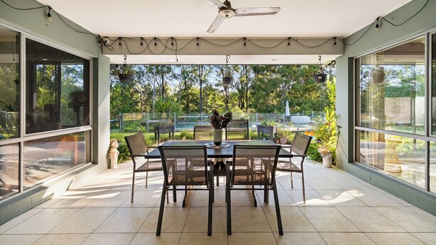 Un patio techado con una mesa y sillas de comedor, un ventilador de techo y grandes ventanas con vistas a un jardín.