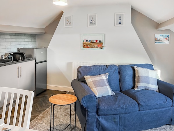 Ein modernes Wohnzimmer mit einem blauen Sofa, einem kleinen Holztisch und einer Küchenzeile mit Kühlschrank und Herd.