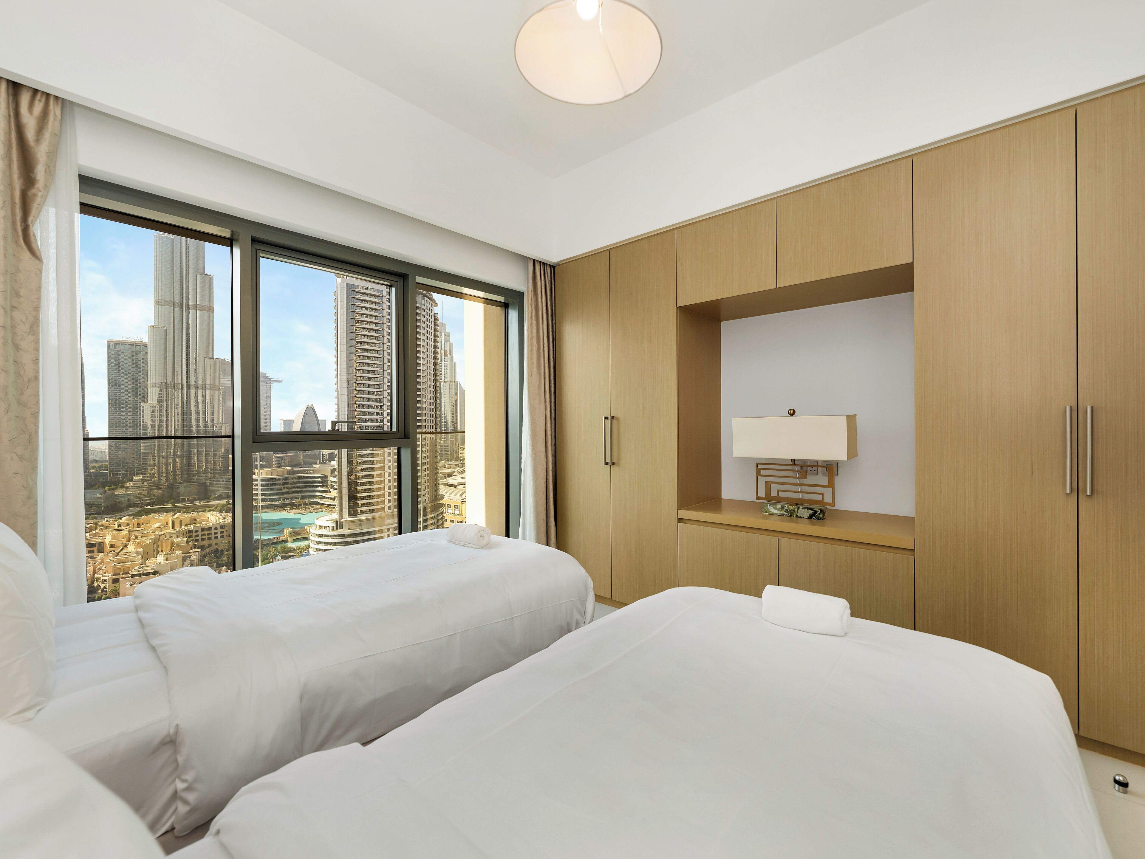 A hotel room with two beds, a large window with a city view, wooden wardrobe, and a bedside table with a lamp.