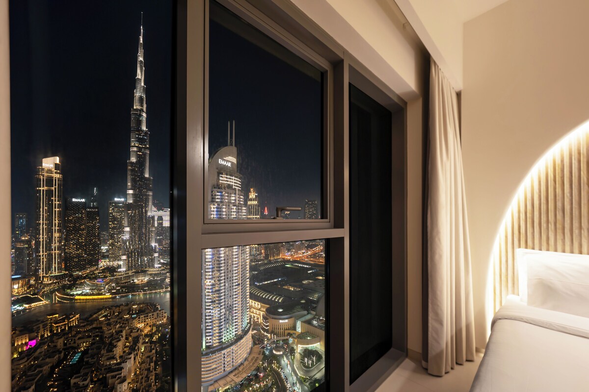 A hotel room with a large window offering a nighttime view of a city skyline, including a prominent skyscraper.