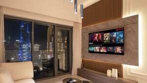 A modern living room with a large TV displaying Netflix content, a city view through floor-to-ceiling windows, and a minimalist design with a glass coffee table and a sofa. - ABI Holidays Burj Royale Fountain Views (Dubai)