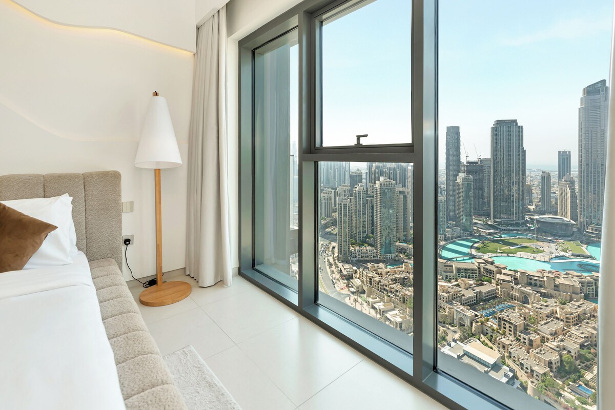 A modern hotel room with a large window offering a city view, a bed with white bedding, and a floor lamp.