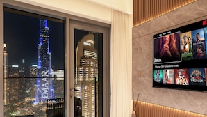A modern living room with a large TV displaying a movie selection, a cityscape view through floor-to-ceiling windows, and a sleek, minimalist design. - ABI Holidays Burj Royale Fountain Views (Dubai)
