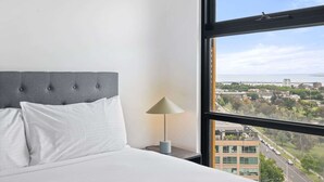 A hotel room with a large window offering a city view, a bed with white linens, and a modern lamp on the nightstand. - Renovated 2-Bed Apartment with Balcony (St Kilda)