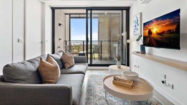 A modern living room with a grey sofa, a round coffee table, and a large TV on the wall. - Renovated 2-Bed Apartment with Balcony (St Kilda)
