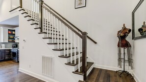 Una escalera con barandillas blancas y peldaños de madera oscura, que conduce a una plataforma con una rejilla de ventilación y un maniquí sobre un pedestal.