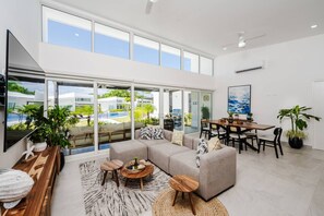 A spacious living area with a large sofa, dining table, and multiple chairs. There are several potted plants and a large window offering a view of a pool and greenery outside. - The Enclave Avellanas - Villa D7 (Playa Avellana)