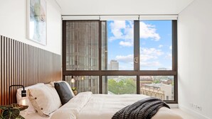 A bedroom with a large window, a bed, a bedside table, and a potted plant. - Moody Luxe Designer 2BR • Dark Oak • Surry Hills (Surry Hills)