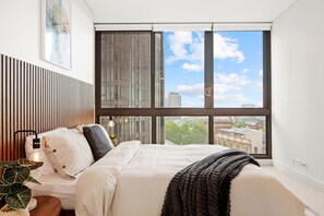 A bedroom with a large window, a bed, a bedside table, and a potted plant. - Moody Luxe Designer 2BR • Dark Oak • Surry Hills (Surry Hills)