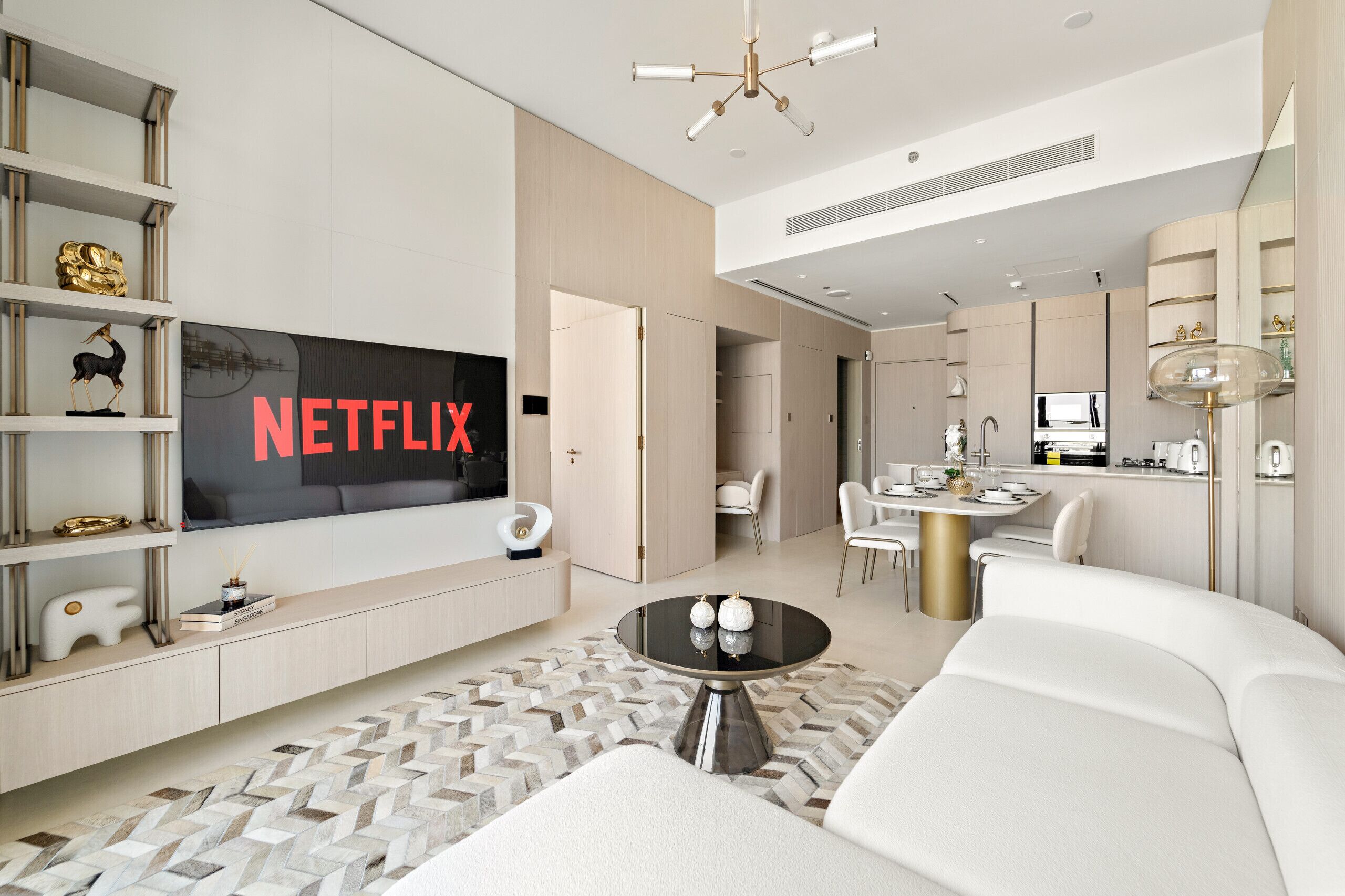 Un salon moderne doté d’un grand téléviseur à écran plat affichant le logo « NETFLIX », d’un coin repas avec une table ronde et d’une cuisine avec un îlot central.