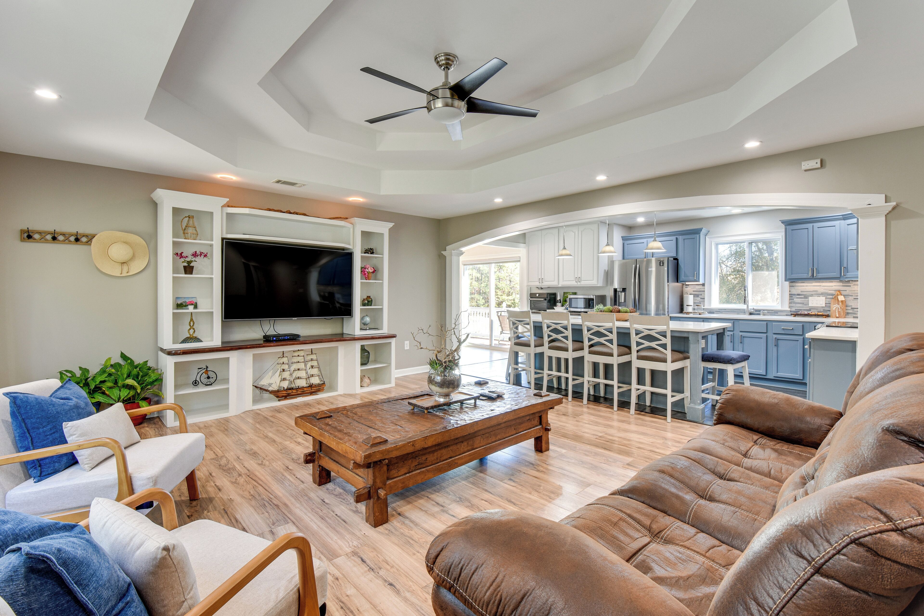 A spacious living room with a large TV, comfortable seating, and a kitchen area visible through an open archway.