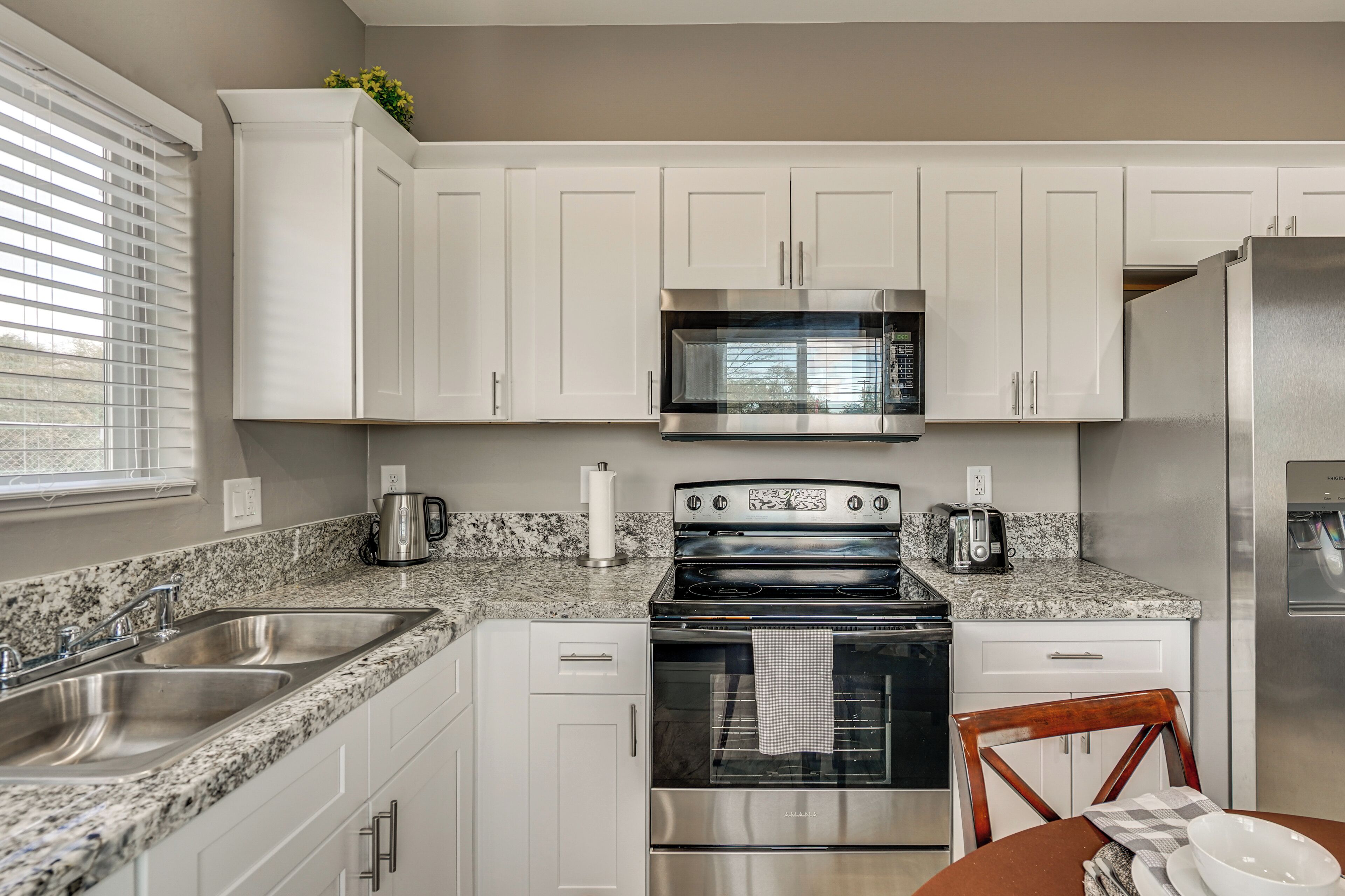 A modern kitchen with white cabinets, granite countertops, stainless steel appliances, and a window with blinds.