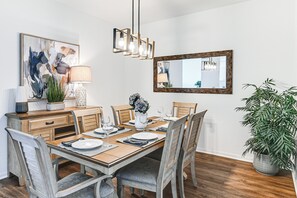 A dining room with a wooden table set for six, surrounded by chairs. There is a chandelier above the table, a mirror on the wall, and a potted plant to the right. - “Overlooking 10th Fairway – 2 Free Rounds of Golf” (Naples)
