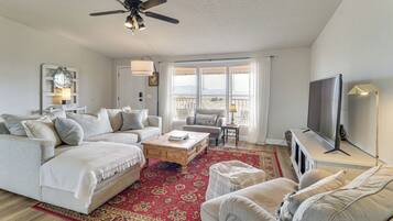 A spacious living room with a large sofa, a coffee table, and a TV. There is a ceiling fan and a window with curtains.