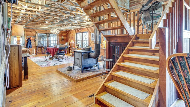 A spacious wooden cabin interior with a central fireplace, a dining area, and a staircase. - 'Mule Creek Lodge' Rural Stay Near Coldwater Lake (Wilmore)
