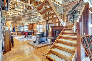A spacious wooden cabin interior with a central fireplace, a dining area, and a staircase. - 'Mule Creek Lodge' Rural Stay Near Coldwater Lake (Wilmore)