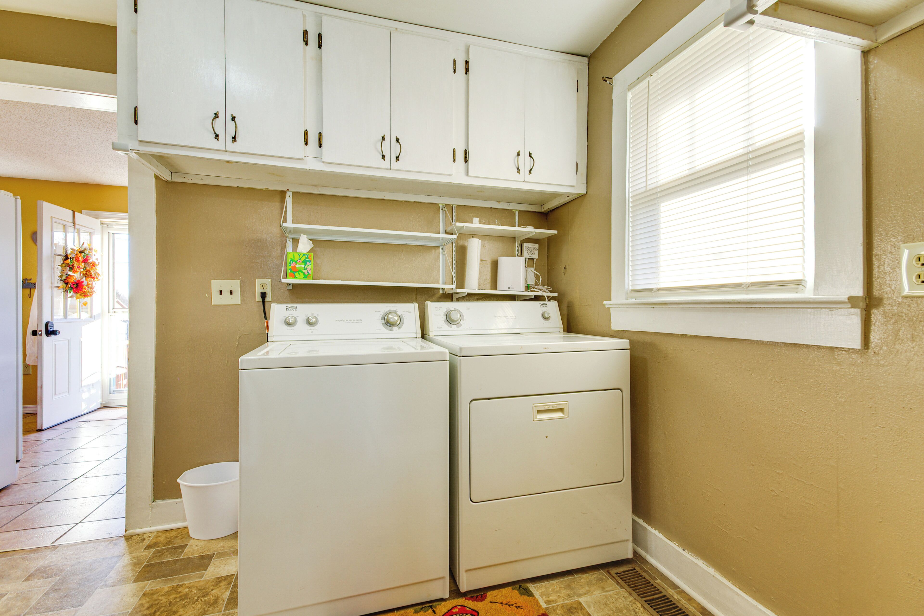 A laundry room with two white washers and dryers, a white cabinet above, and a window with blinds.