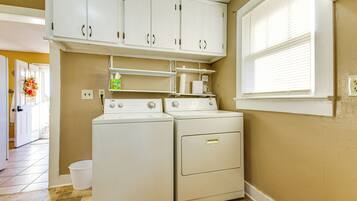 A laundry room with two white washers and dryers, a white cabinet above, and a window with blinds.