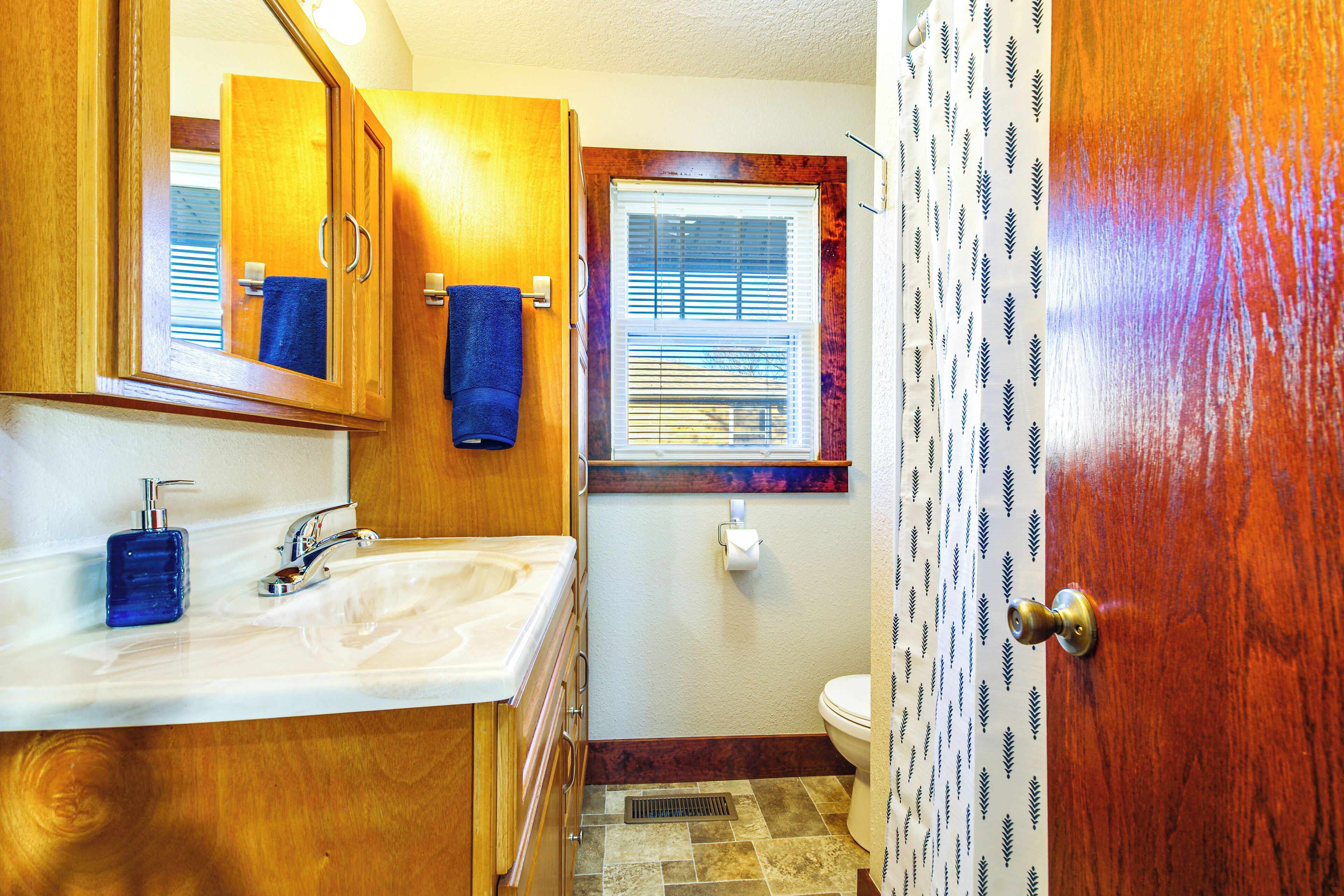 Un baño con armarios de madera, un lavabo blanco, una toalla azul, una ventana con persianas y una cortina de ducha estampada.