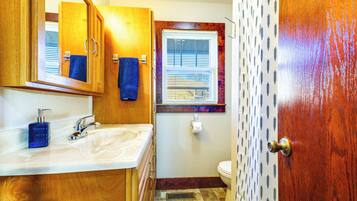 Un baño con armarios de madera, un lavabo blanco, una toalla azul, una ventana con persianas y una cortina de ducha estampada.
