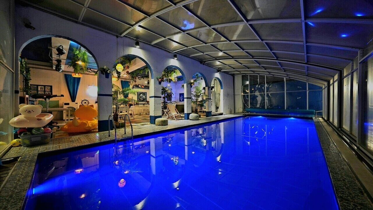 An indoor swimming pool with a blue-lit floor, arched ceilings, and various plants and decorations.