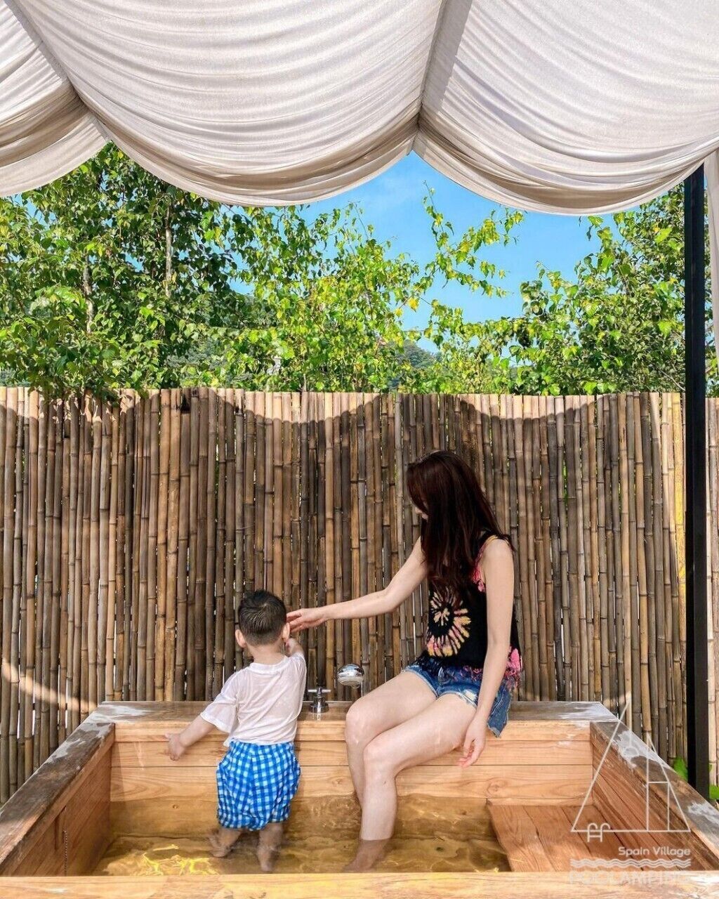 A wooden hot tub with a bamboo backdrop, a child and an adult interacting, and a clear blue sky above.