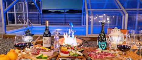 An outdoor dining setup with a fire pit, various meats, cheeses, and drinks on a wooden table, set against a night sky and glass structures.