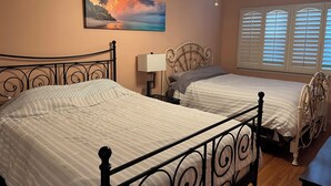 Een slaapkamer met twee bedden, een plafondventilator, een schilderij van een strandgezicht en een raam met jaloezieën.