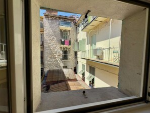 Blick aus einem Fenster eines Gebäudes mit einer steinernen Fassade, Balkonen mit Geländern und einer kleinen Außenfläche mit einer Holzterrasse.