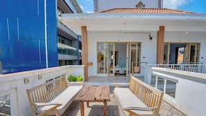 Un balcon doté de mobilier en bois, d’un sol carrelé et offrant une vue sur un immeuble à la façade bleue.